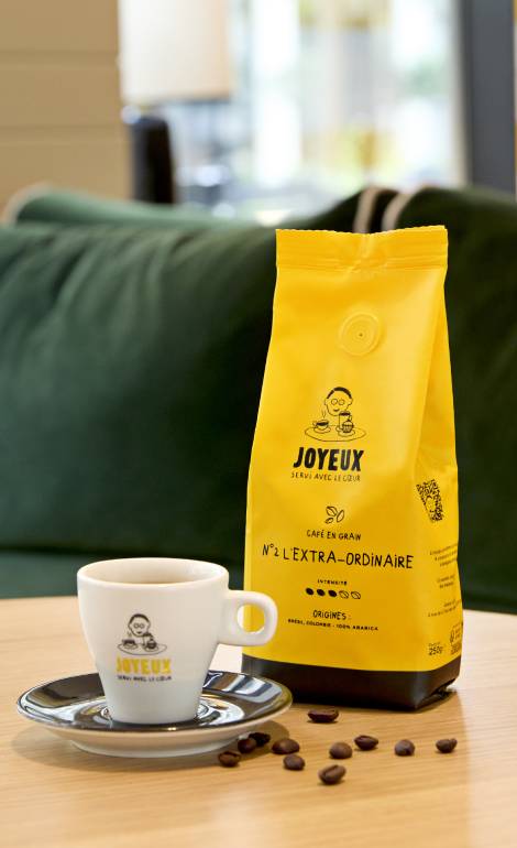 Visuel d'un paquet de café L'Extra-ordinaire de Café Joyeux, une tasse de café avec le logo Café Joyeux et des grains de café posée sur une table.