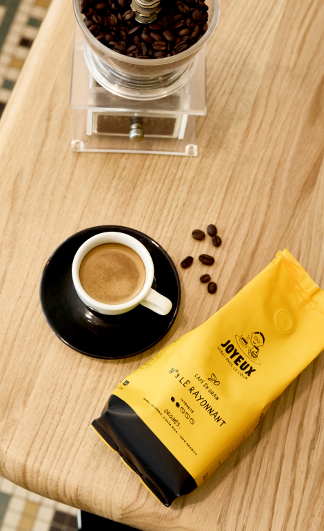 Photo prise au survol d'un paquet de café en grain Le Rayonnant de Café Joyeux couché sur une table, avec une tasse de café, des grains de café et une machine à moulin à café